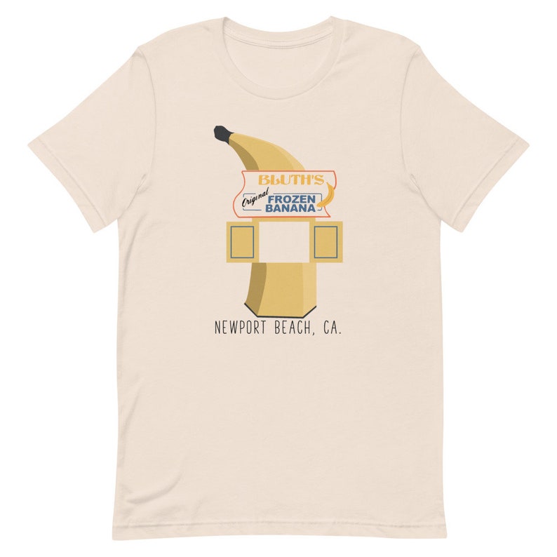 May include: A beige t-shirt featuring a graphic of a yellow banana and a sign that reads "Bluth's Original Frozen Banana" with the text "Newport Beach, CA."