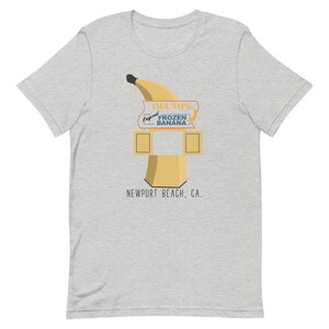May include: A grey t-shirt featuring a graphic of a yellow banana and a sign that reads "Bluth's Original Frozen Banana" and "Newport Beach, CA."