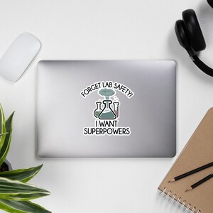 May include: A white sticker featuring the text "FORGET LAB SAFETY! I WANT SUPERPOWERS" and lab equipment illustrations. The sticker has a cartoon style and a science theme. The sticker is on a laptop.