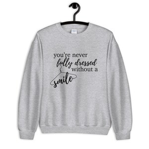 Puede incluir: Una sudadera gris con el texto "you're never fully dressed without a smile" impreso en la parte delantera. El texto está en negro y presenta una percha estilizada con la palabra "smile" escrita en ella.