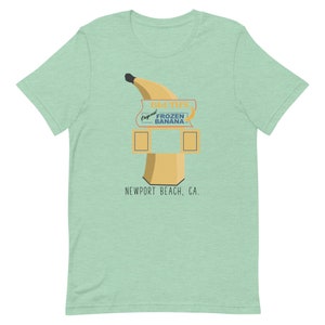 May include: A green t-shirt featuring a cartoon illustration of a banana stand. The stand has a sign that reads "Original Frozen Banana" and "D.U.T.S." The stand is located in Newport Beach, CA.