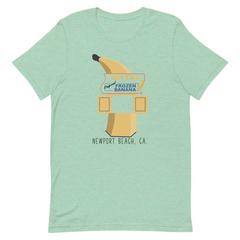 May include: A green t-shirt featuring a cartoon illustration of a banana stand. The stand has a sign that reads "Original Frozen Banana" and "D.U.T.S." The stand is located in Newport Beach, CA.