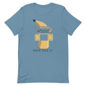 May include: A light blue t-shirt featuring a graphic of a yellow banana and a sign that reads "Bluth's Original Frozen Banana, Newport Beach, CA."