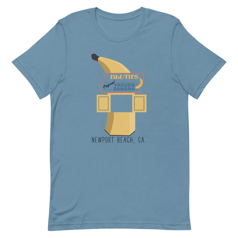 May include: A light blue t-shirt featuring a graphic of a yellow banana and a sign that reads "Bluth's Original Frozen Banana, Newport Beach, CA."