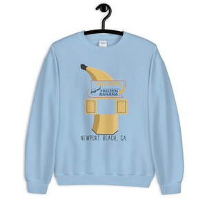 May include: Light blue sweatshirt with a graphic of a banana-shaped frozen banana stand with the text "Doug's Original Frozen Banana" and "Newport Beach, CA."