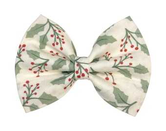 May include: A cream-colored hair bow with a festive holly berry pattern. The bow features green leaves and red berries, creating a seasonal design. The bow is tied in the center, with two loops on each side.