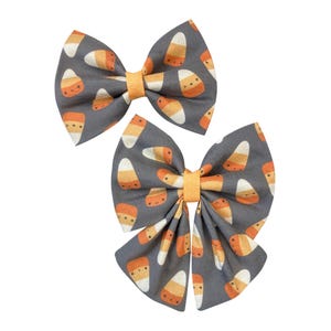 May include: Two hair bows featuring a dark grey fabric base with a repeating pattern of orange, yellow, and white candy corn shapes. Each bow is centred with a solid orange ribbon band, creating a festive look with a classic bow silhouette.