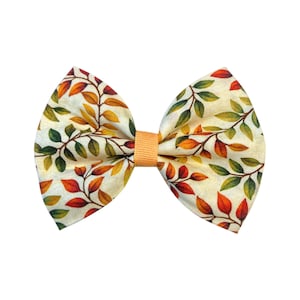 May include: A fabric bow tie featuring a pattern of autumn leaves in orange, yellow, and green on an off-white backdrop. The centre of the bow is a solid peach colour.