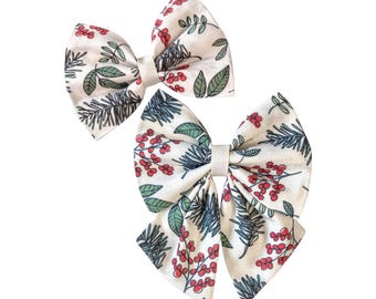 May include: Two hair bows featuring a cream-colored fabric with a repeating pattern of red berries, green leaves, and dark pine needles. Each bow is secured in the center with a matching cream-colored fabric band, creating a festive botanical design.