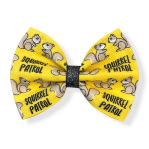 May include: A yellow bow tie with a repeating pattern of cartoon squirrels and the words "SQUIRREL PATROL" in black. The center of the bow tie is a black glittery band.