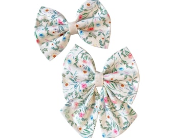 May include: Two hair bows made of off-white fabric featuring a delicate floral pattern with green leaves, pink, blue, and yellow flowers. Each bow is gathered in the center with a matching off-white fabric band, creating a classic bow shape.