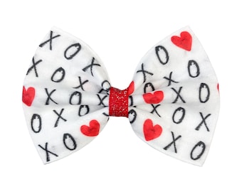 May include: White fabric bow with a red glitter center. The bow is covered in a pattern of black Xs and Os and red hearts.