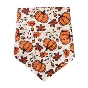 A dog bandana with fall pumpkins and leaves scattered around a beige background.