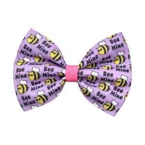 May include: A lavender bow tie with a pink centre. The bow tie features a pattern of yellow and black cartoon bees and the words "Bee Mine". Suitable for pets.