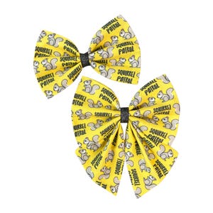 May include: Two yellow hair bows featuring a repeating pattern of grey squirrels and the black text "SQUIRREL PATROL". Each bow is gathered in the center with a thin, black, glittery fabric band, creating a structured, multi-layered appearance.