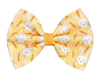 May include: A yellow bow with a white bunny and carrot pattern. The bow is tied with a yellow ribbon.