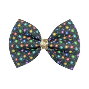 May include: A fabric bow with a festive design. The bow features a dark background adorned with colorful string lights and small white stars. The center knot is gold glitter. This hair accessory is perfect for the holidays.