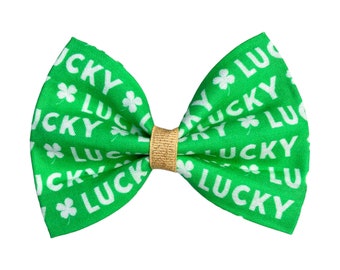 May include: A green fabric bow tie with a gold center. The bow tie is covered in a repeating pattern of white shamrocks and the word "lucky".