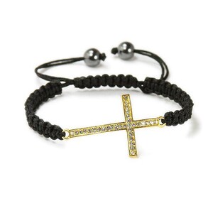 May include: A black braided bracelet with a gold-colored cross pendant. The cross is encrusted with small, clear crystals. The bracelet has adjustable black cords and two small, dark gray beads.