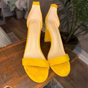 Puede incluir: Un par de sandalias abiertas de color amarillo brillante con tacón de bloque y correa al tobillo. La plantilla interior de cada zapato presenta el nombre de la marca ILLUDE impreso en un color claro. El material exterior tiene una textura mate.
