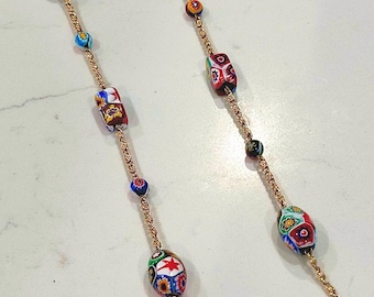 May include: A gold-toned necklace featuring colorful, patterned glass beads. The beads are various shapes and sizes, with intricate designs in red, blue, green, and yellow. The necklace is displayed on a white surface.