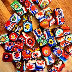 May include: A collection of colorful, rectangular glass beads with floral and geometric patterns. The beads feature red, blue, green, and yellow designs, with white and black accents. The beads are scattered on a wooden surface.