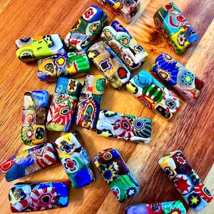 May include: A collection of colorful, rectangular glass beads with intricate floral and geometric patterns. The beads feature a variety of colors, including red, blue, green, yellow, and white. The beads are arranged on a wooden surface.