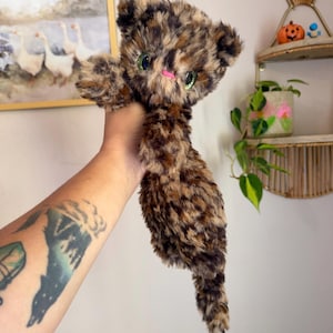 May include: A plush toy cat with long, soft faux fur in a mottled tortoiseshell pattern of black, brown, and tan. The stuffed animal features large, bright green plastic eyes and a small, stitched pink nose, with a long tail and soft paws.