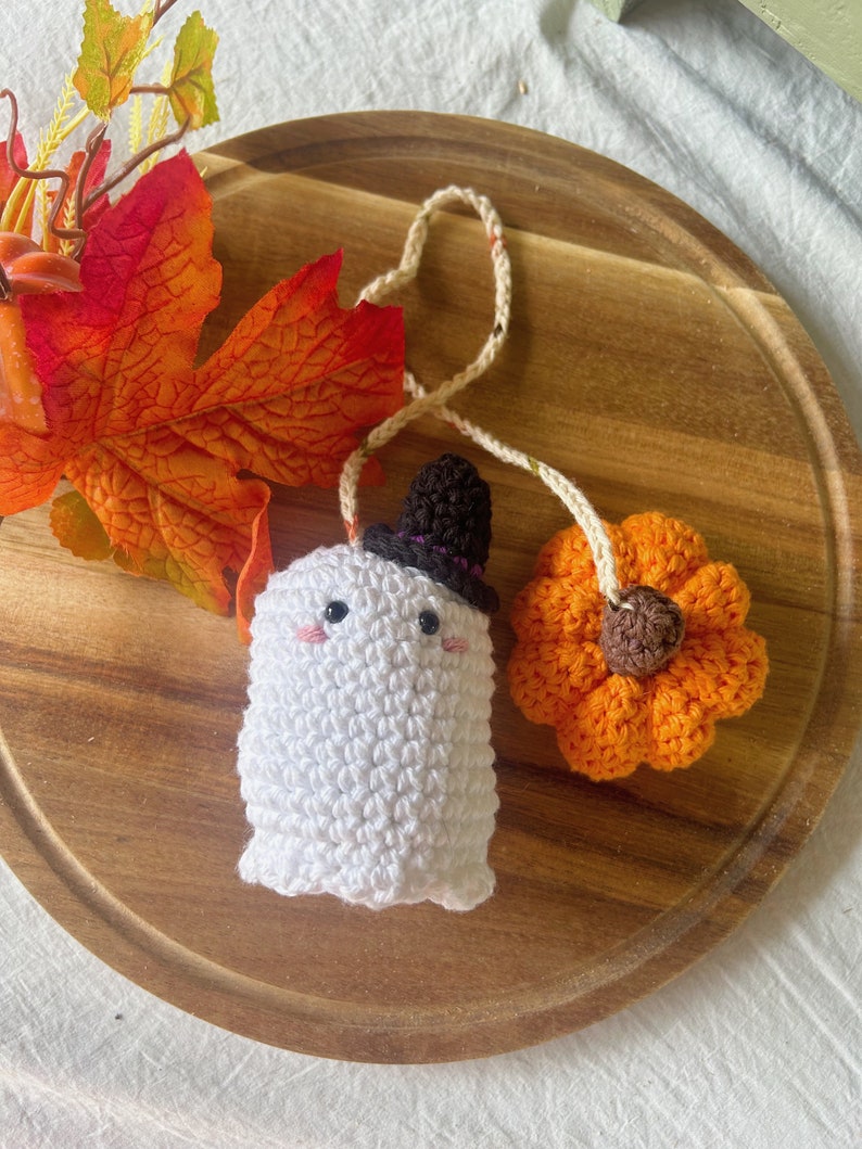 Puede incluir: Un fantasma de crochet con un sombrero de copa negro y una calabaza naranja de crochet. El fantasma y la calabaza est&aacute;n conectados por una cuerda blanca.