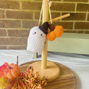 Puede incluir: Un fantasma blanco de crochet con un sombrero de bruja negro y una calabaza naranja de crochet colgando de un soporte de madera. El soporte est&aacute; sobre una superficie de madera con follaje de oto&ntilde;o.