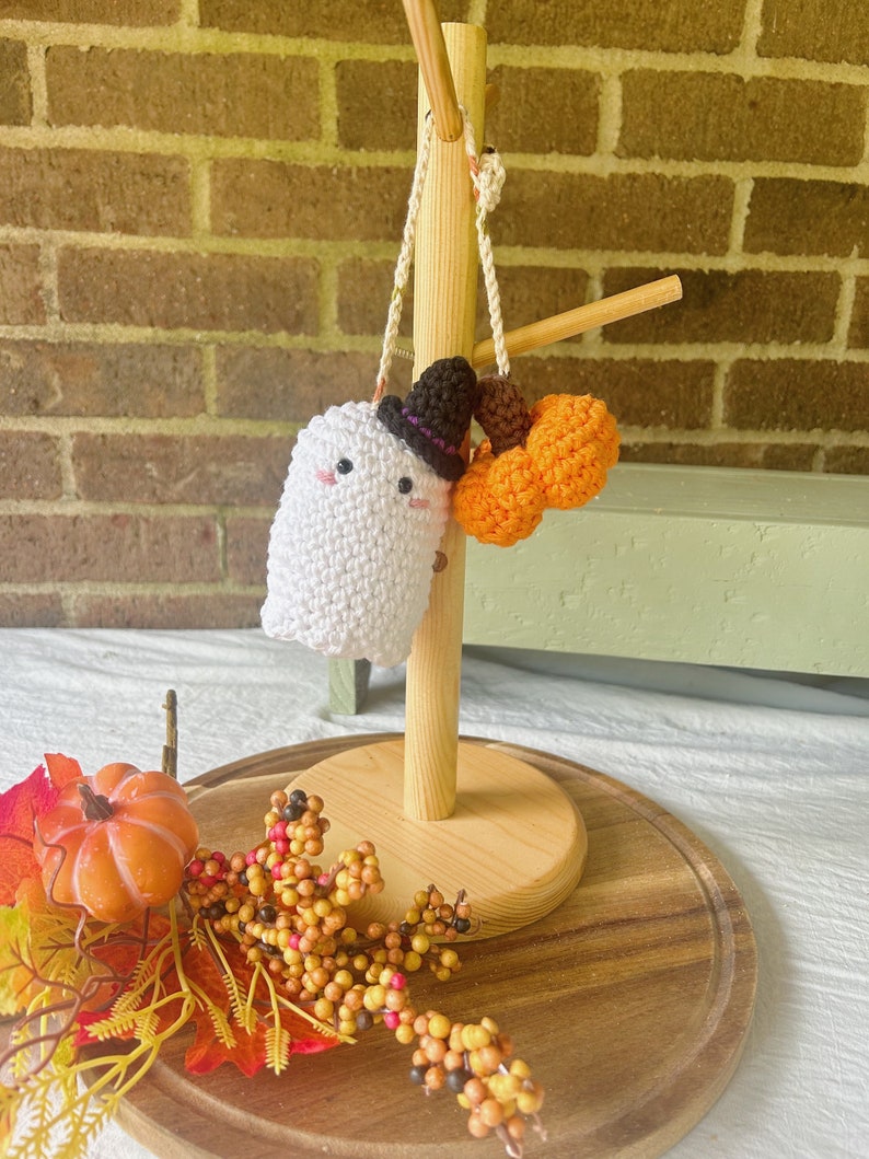 Puede incluir: Un fantasma blanco de crochet con un sombrero de bruja negro y una calabaza naranja de crochet colgando de un soporte de madera. El soporte est&aacute; sobre una superficie de madera con follaje de oto&ntilde;o.
