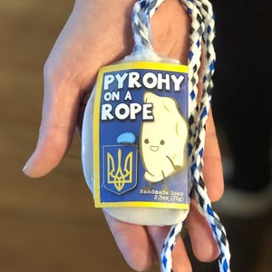 May include: A white handmade soap shaped like a pierogi with a blue and yellow Ukrainian flag and the text "PYROHY ON A ROPE" on a blue and white rope.