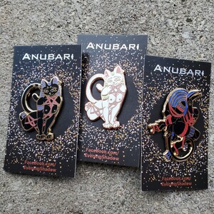 Könnte beinhalten: Drei schwarze Emaille-Pins mit goldenen Akzenten, die eine Katze in verschiedenen Posen zeigen. Jeder Pin trägt den Text "Anubari" und eine Website-Adresse. Die Katze trägt ein rot-pinkes Geschirr.