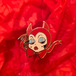 Puede incluir: Un pin de esmalte rojo y dorado con una mujer diablo de dibujos animados con ojos azules, cuernos rojos y una pajarita roja. El pin tiene un contorno dorado sobre un fondo rojo.
