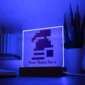Op de afbeelding: Een blauw LED-licht met een gepixelde afbeelding van een videogamepersonage. Het licht heeft een houten basis en de tekst "Your Name Here" is op de voorkant gedrukt.