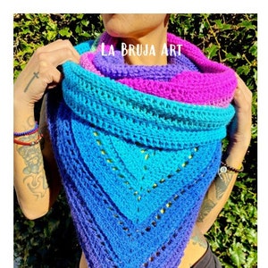 May include: A colorful crocheted shawl in shades of purple, pink, blue, and turquoise. The shawl is triangular in shape and has a lacy pattern. The text "LA BRUJA ART" is visible on the shawl.