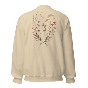 May include: A cream-coloured crewneck sweatshirt featuring a botanical illustration on the back. The design includes two symmetrical floral branches, two butterflies, and a crescent moon. The text "WALKING BESIDE YOU" is printed below the graphic.