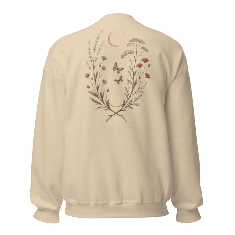 May include: A cream-coloured crewneck sweatshirt featuring a botanical illustration on the back. The design includes two symmetrical floral branches, two butterflies, and a crescent moon. The text "WALKING BESIDE YOU" is printed below the graphic.