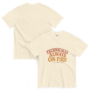 Puede incluir: Una camiseta de manga corta color crema con el texto "TECHNICALLY ALWAYS ON FIRE" en naranja y marr&oacute;n, un peque&ntilde;o icono de llama sobre la palabra "ON" y el texto "it's a CRPS thing" impreso en letras m&aacute;s peque&ntilde;as debajo.
