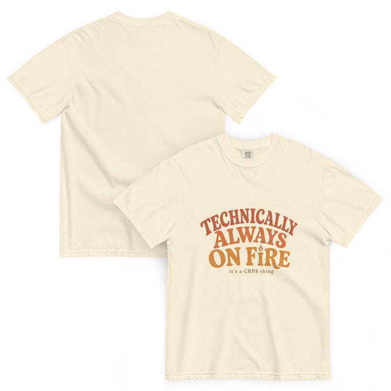 Puede incluir: Una camiseta de manga corta color crema con el texto "TECHNICALLY ALWAYS ON FIRE" en naranja y marr&oacute;n, un peque&ntilde;o icono de llama sobre la palabra "ON" y el texto "it's a CRPS thing" impreso en letras m&aacute;s peque&ntilde;as debajo.