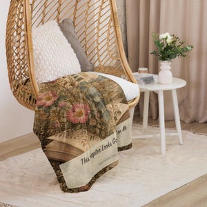 K&ouml;nnte beinhalten: Eine weiche Wohndecke mit einem botanischen Druck im Vintage-Stil, der offene B&uuml;cher, rosa Blumen und gr&uuml;nes Blattwerk zeigt. Der Text "This Chapter Looks Good On Me" ist in schwarzer Schrift auf einem hellen Banner auf dem Stoff gedruckt.