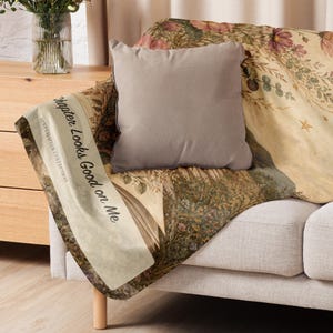 K&ouml;nnte beinhalten: Ein einfarbiges, taupefarbenes quadratisches Dekokissen auf einer weichen Decke mit einer botanischen Vintage-Illustration aus Blumen, Bl&auml;ttern und Sternen. Die Decke zeigt den Text "Chapter Looks Good on Me" und "NEXT CHAPTER COLLECTION".