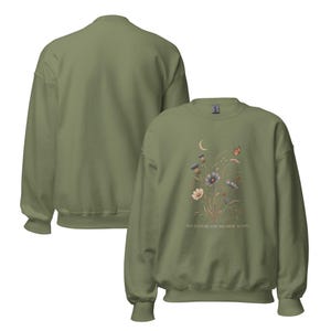 May include: An olive green crewneck sweatshirt featuring a graphic print of wildflowers, a crescent moon, and a butterfly. The text ADVENTURE FOUND HER AGAIN is printed below the floral design. The sweatshirt has long sleeves and ribbed cuffs.