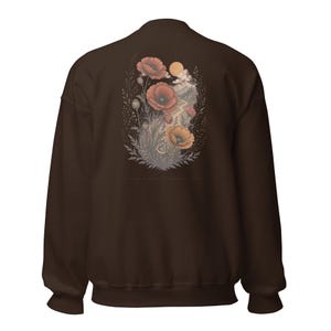 May include: A dark brown crewneck sweatshirt featuring a back graphic of red and orange poppies, foliage, a mountain landscape, and a sun. The design includes a small crescent moon and faint text at the bottom, showcasing a detailed botanical illustration.