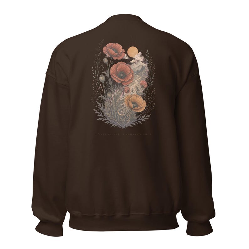 May include: A dark brown crewneck sweatshirt featuring a back graphic of red and orange poppies, foliage, a mountain landscape, and a sun. The design includes a small crescent moon and faint text at the bottom, showcasing a detailed botanical illustration.