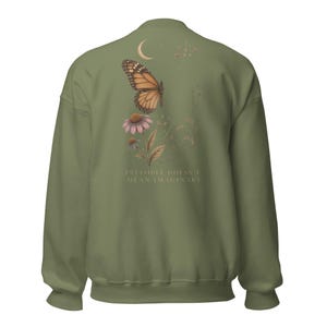 May include: An olive green crewneck sweatshirt featuring a back graphic of a monarch butterfly, a pink coneflower, and a crescent moon. The text "INVISIBLE DOESN'T MEAN IMAGINARY" is printed in a light color below the floral and insect illustration.