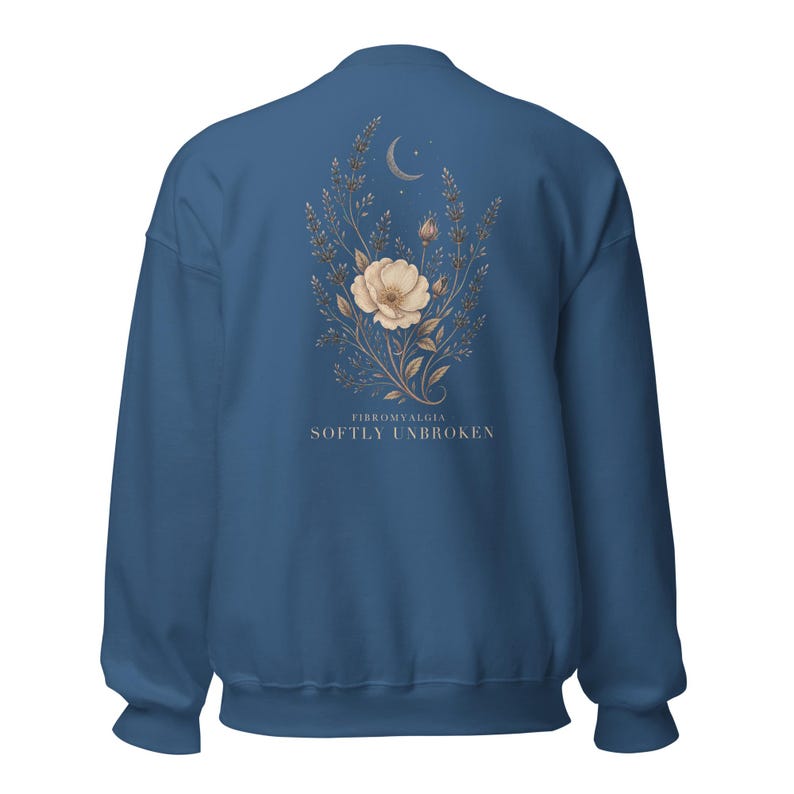 May include: A dark blue crewneck sweatshirt featuring a central graphic on the back. The design displays a beige floral arrangement with a crescent moon and stars above it. Below the flowers, the text reads FIBROMYALGIA SOFTLY UNBROKEN.