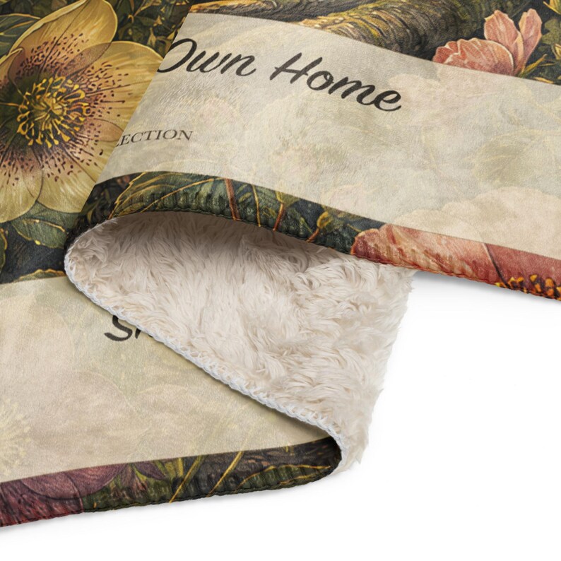 May include: A folded throw blanket with a cream-colored faux sherpa underside and a printed top featuring botanical illustrations of flowers and leaves in shades of yellow, pink, and green. The text "Own Home" and "ECTION" are visible on the print.