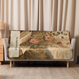 K&ouml;nnte beinhalten: Eine rechteckige Wohndecke auf einem Sofa mit einer Illustration im Vintage-Stil, die aufgeschlagene Buchseiten und ein &uuml;ppiges Blumenarrangement mit rosa Bl&uuml;ten und gr&uuml;nem Blattwerk zeigt. Der Text "This Chapter Looks Good on Me" ist auf der linken Seite aufgedruckt.