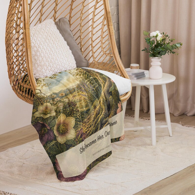 May include: A rectangular throw blanket featuring a detailed botanical illustration of hellebore flowers and tree roots. The text "She came Her Own Home" and "NEXT CHAPTER COLLECTION" is printed in black on a light cream-colored rectangular banner.